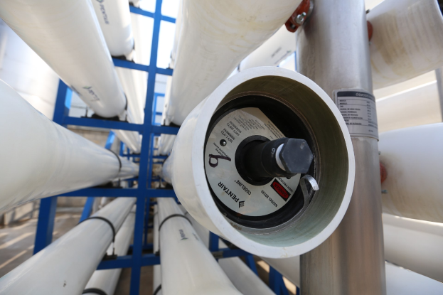 Nahaufnahme eines Metallrohrsystems in einem Gebäude, das einen Metallstab (wahrscheinlich Teil eines Wasserheizers) mit angeschlossenen Rohren und eine Wand im Hintergrund zeigt.
