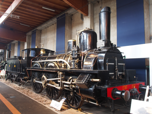 Eine alte Dampflokomotive in einem Museums-Setting, umgeben von Eisenbahnschienen, Informations-Tafeln und anderen Ausstellungsstücken, mit einer beleuchteten Decke und Wand im Hintergrund.