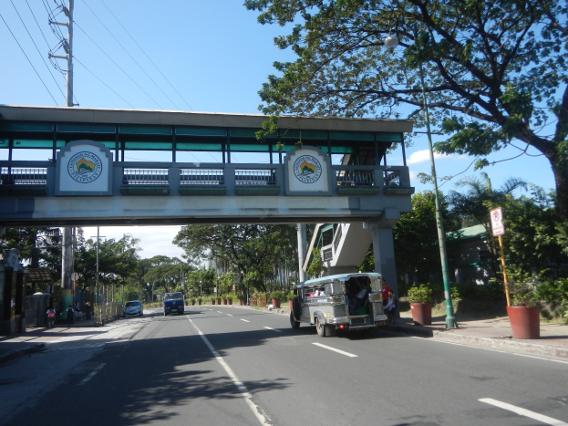 Ein Bus fährt eine von Bäumen gesäumte Straße mit einer Brücke im Hintergrund, mit Text- und Logo-Schildern, Strommasten mit Drähten, Laternen und ein paar Fußgängern auf dem Gehweg unter einem bewölkten Himmel.
