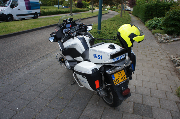 Ein auf der Straße geparktes Polizeimotorrad mit einem Helm darauf, mit Bäumen, Pfählen, Gebäuden, Pflanzen, Gras, Steinen und anderen Fahrzeugen im Hintergrund.