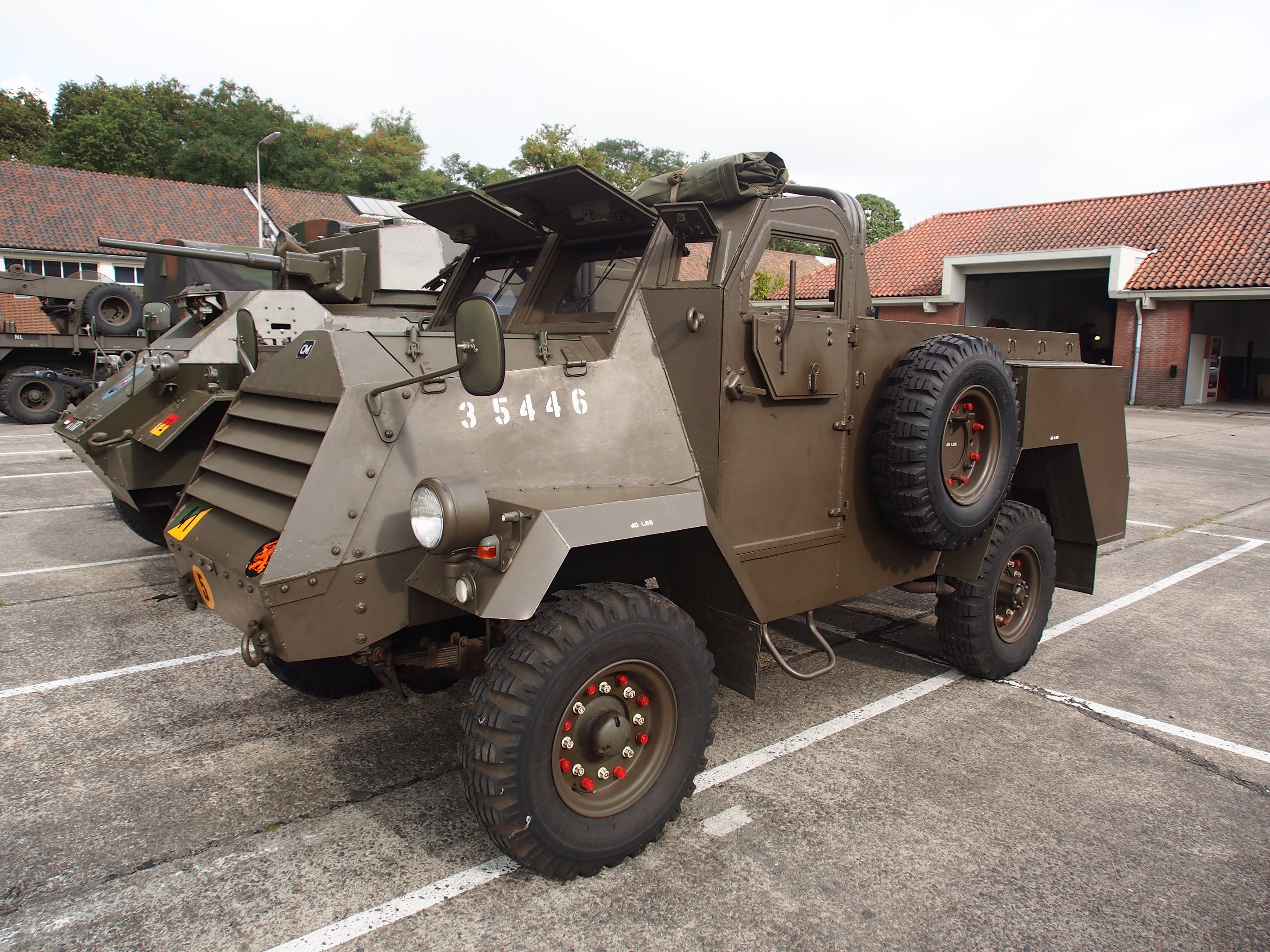 Militärischer gepanzerter Mannschaftstransporter mit einer montierten großen Kanone auf einem Parkplatz neben einem Gebäude geparkt, umgeben von Bäumen unter einem klaren blauen Himmel.