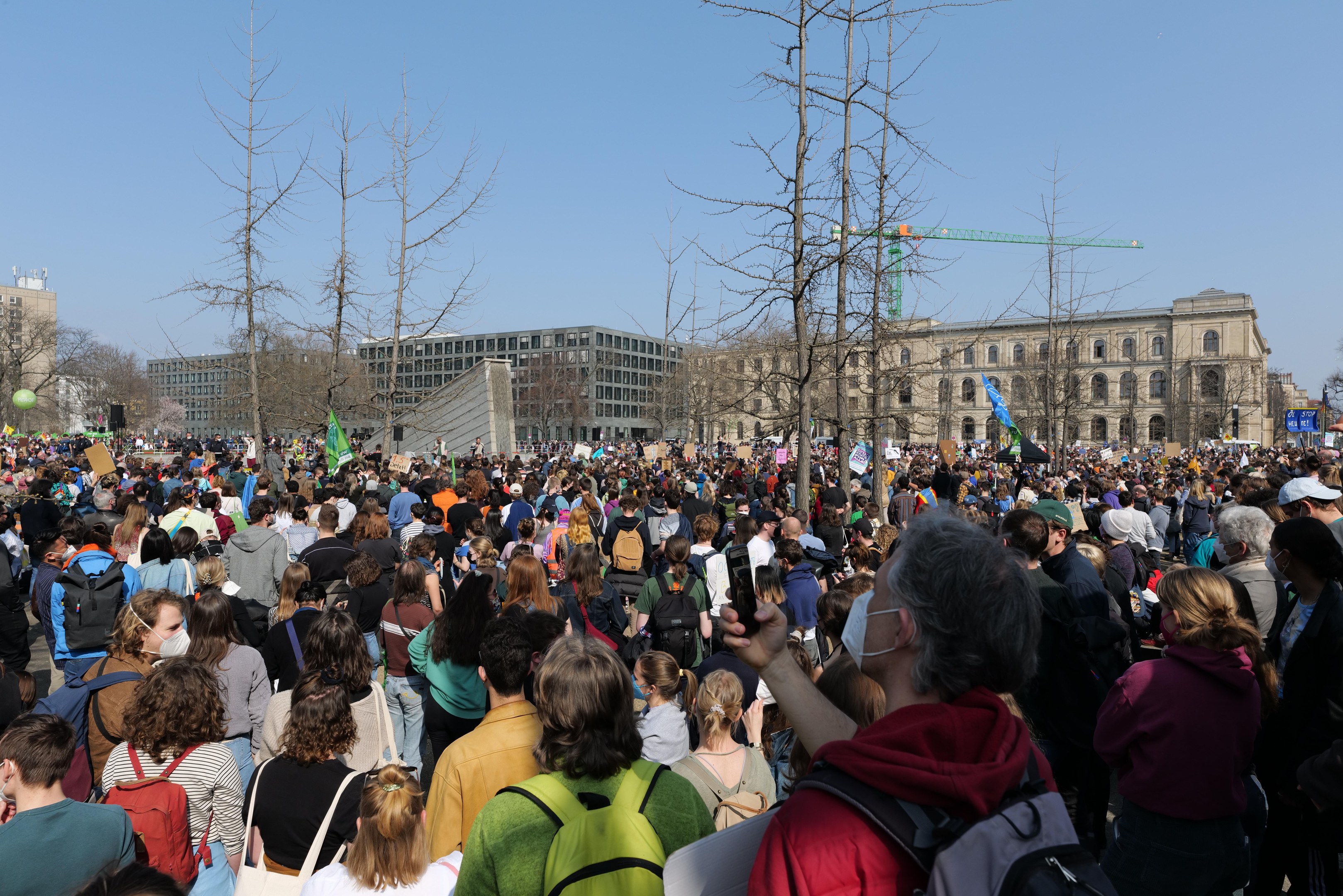 Eine große Menge Menschen nimmt an einer Klimaprotestaktion vor einem Gebäude mit Bäumen und einem klaren Himmel teil.