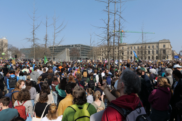 Eine große Menge Menschen nimmt an einer Klimaprotestaktion vor einem Gebäude mit Bäumen und einem klaren Himmel teil.