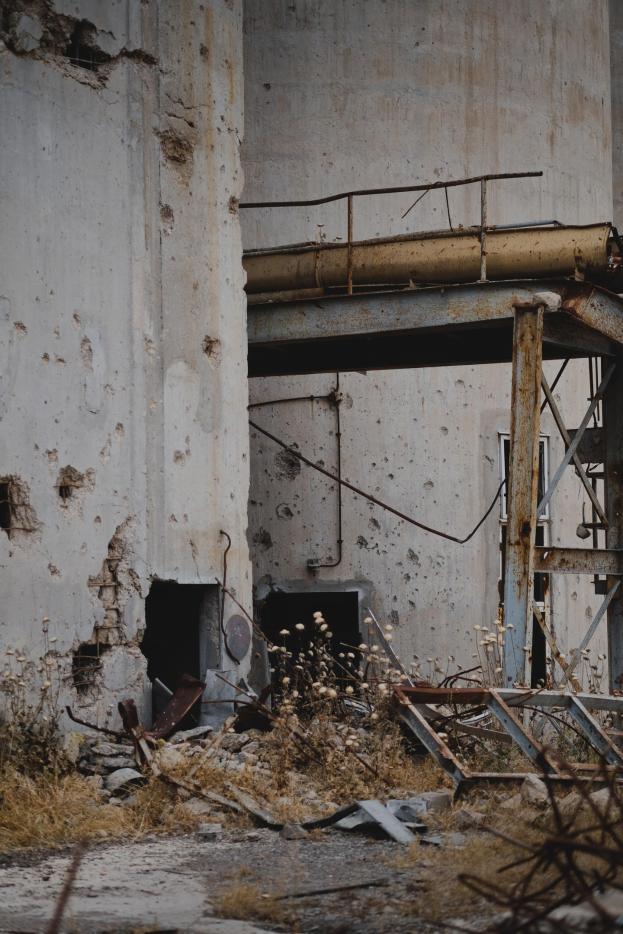 Altes Gebäude mit bröckelnden Wänden, zerbrochenen Fenstern und einem zentralen Metallgerüst, umgeben von Pflanzen, Eisenstangen und verstreutem Schutt.