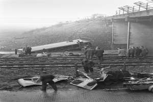 Ein Schwarz-Weiß-Bild einer Zugkatastrophe auf den Schienen, mit einer Gruppe von Menschen um das Wrack herumstehend, einem Fahrzeug im Vordergrund und einer Brücke, Polen, Drähte und Himmel im Hintergrund.