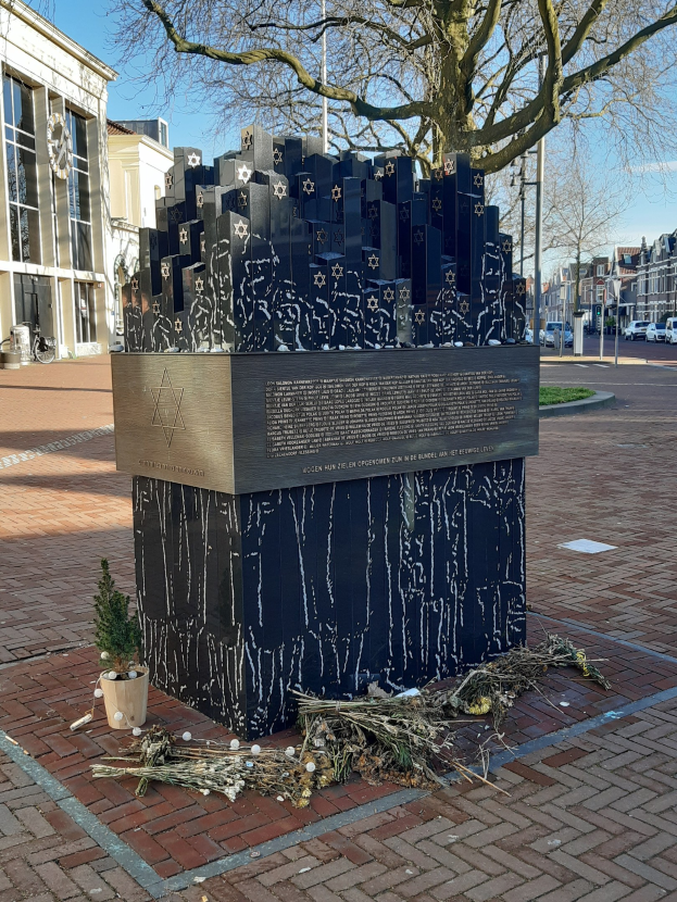 Denkmal in einem Stadtplatz, das Holocaust-Opfern gewidmet ist, mit einem Baum davor, einer Topfpflanze, verstreuten trockenen Blättern und im Hintergrund Gebäude, Fahrzeuge, Masten und Bäume.
