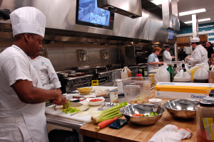 Eine Gruppe von Köchen in einer Küche, die Essen auf einem Schneidbrett zubereiten, mit einem Koch im Vordergrund in einer weißen Schürze und Mütze, der ein Messer hält und von Utensilien, Schüsseln, Gemüse, einem Handy, Flaschen und anderen Gegenständen umgeben ist, mit einem Fernseher und Deckenlampen im Hintergrund.