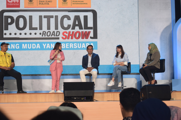 Eine Gruppe von Menschen sitzt auf einer Bühne mit einer Frau in der Mitte, die ein Mikrofon hält, eine Zuschauermenge im Vordergrund und ein Banner mit der Aufschrift "Politischer Roadshow 2019" im Hintergrund.