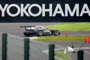 Schwarzer Sportwagen auf einer Rennstrecke mit Verkehrszeichen, einem "Yokohama"-Schild im Hintergrund, Gras, einem Zaun und einem Lautsprecher im Vordergrund.