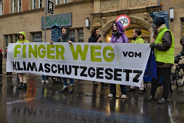 Eine Gruppe von Menschen steht auf der Straße und hält ein Schild hoch, auf dem "Finger Weg Klimaschutzgesetz" steht, mit einem Gebäude, einem Schild und einem Fahrrad im Hintergrund.
