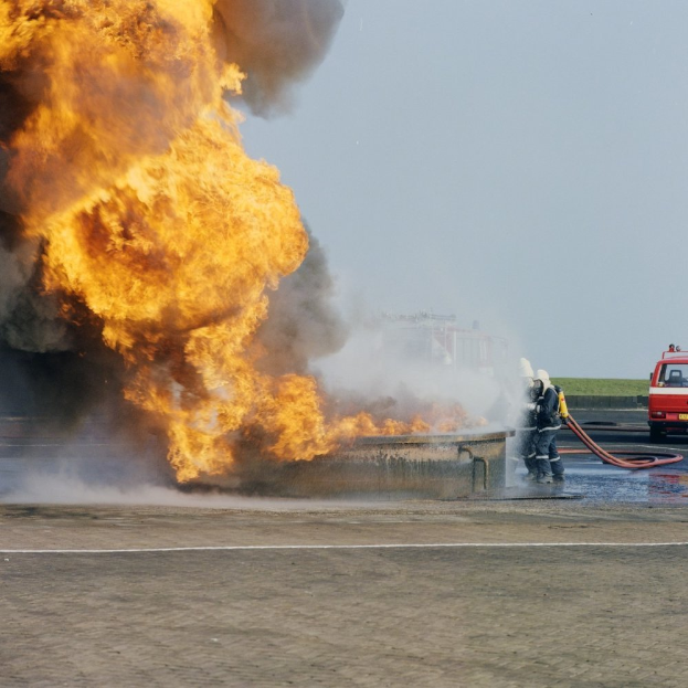 Ein Feuerwehrauto steht in Flammen am Rande einer Straße, zwei Personen mit Helmen und Schlöschen in der Nähe und ein Fahrzeug sowie der Himmel im Hintergrund.