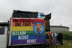 Eine Gruppe von Menschen steht vor einem Lkw mit einer Fahne, auf der "Ein Kampf, ein Kampf, zurückerobern, widerstehen, revoltieren" steht, der auf einem grasigen Bereich mit Pflanzen und einem Gebäude im Hintergrund unter einem klaren blauen Himmel geparkt ist.