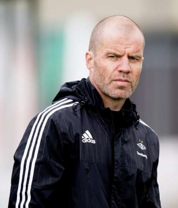 Baldiger Trainer in marineblauer Jacke mit weißen Streifen und weißem Kragen, der auf einem Fußballfeld steht und die Hände gefaltet hat