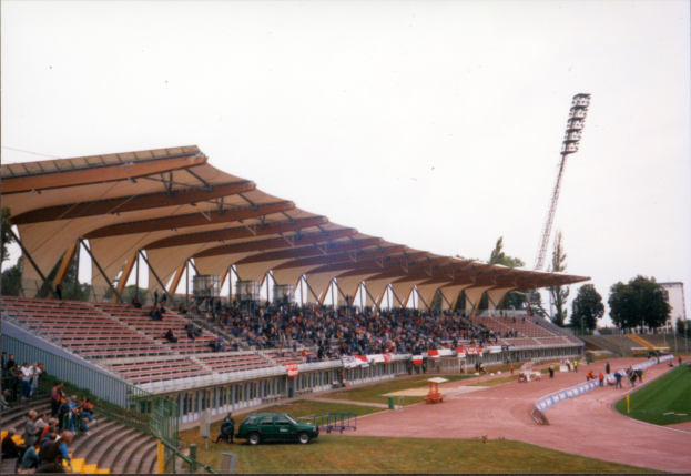 Altes Schwarz-Weiß-Foto eines Stadions voller Zuschauer, das sitzende Fans unter dem Dach, ein geparktes Auto, stehende Besucher auf dem Feld, Stützstangen, einen Metallrahmen, einen umgebenden Zaun, eine Straßenlaterne, Bäume, eine ferne Turm und einen bewölkten Himmel zeigt.