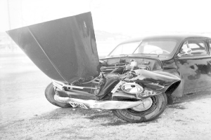 Ein stark beschädigtes Schwarz-Weiß-Foto eines bei einem Crash beteiligten Autos mit eingedellter Motorhaube, zersplitterten Fenstern und einer offenen Fahrertür, die den Motor zeigt.