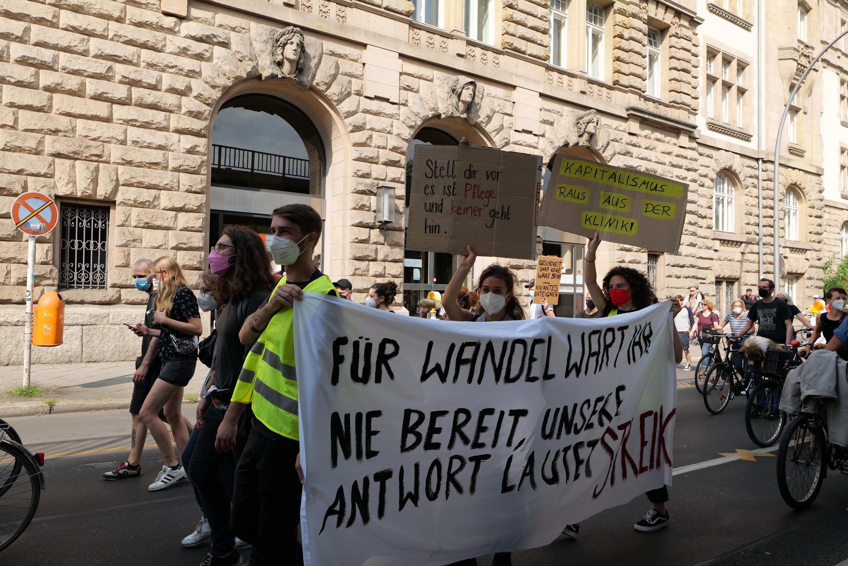 Eine Gruppe von Menschen marschiert auf einer Stadtstraße bei einer Demonstration, einige halten Schilder und andere fahren Fahrräder, im Hintergrund ein historisches Gebäude mit Rundbogenfenstern, Säulen und Skulpturen.