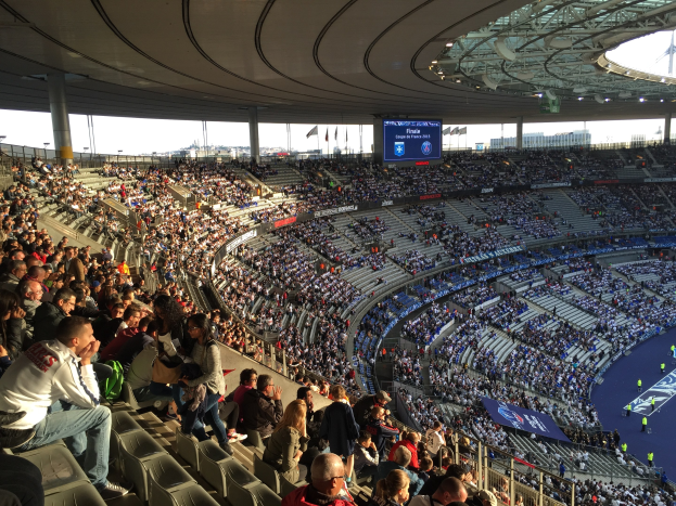 Eine große Menschenmenge sitzt in einem Stadion und schaut ein Fußballspiel. Rechts im Bild befindet sich eine Bühne mit ein paar Menschen darauf und im Hintergrund sind Fahnen, Stangen und ein Bildschirm zu sehen. Der Himmel ist oben im Bild zu sehen und das Stadion scheint die Allianz Arena in München, Deutschland zu sein.