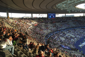 Große Menschenmenge in einem Stadion bei einem Fußballspiel mit einer Bühne, Fahnen, Stangen, einem Bildschirm und dem Allianz Arena in München, Deutschland im Hintergrund.