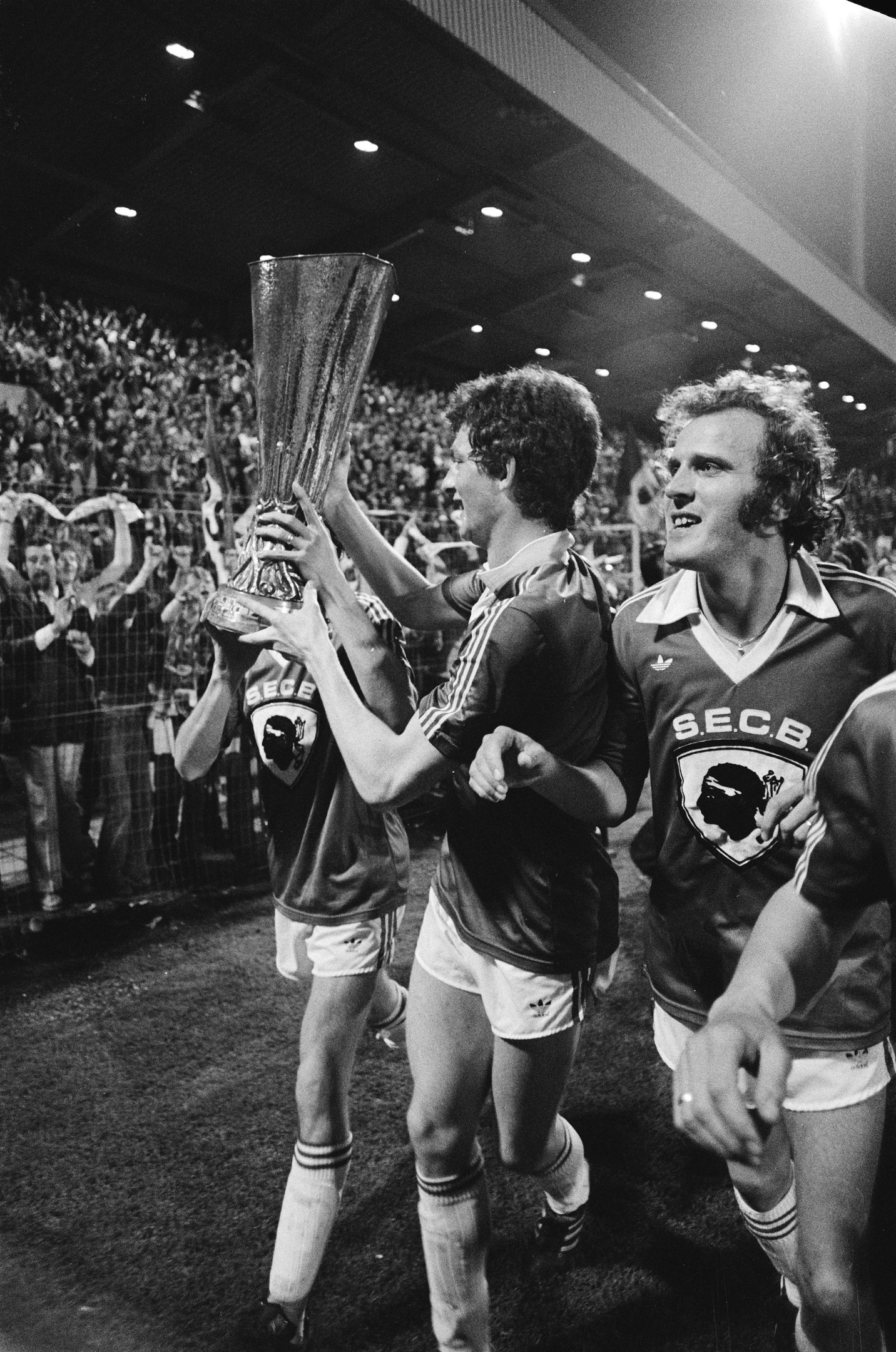 Schwarzes und weißes Foto von Fußballspielern, die mit einem Pokal auf einem Feld feiern, umgeben von einer jubelnden Menge hinter einem Zaun unter Stadionlichtern.