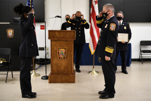 Gruppe uniformierter Personen in Masken vor einem Rednerpult mit Mikrofon, umgeben von Flaggen und Möbeln, während einer Vereidungszeremonie, bei der eine Person ein Zertifikat hält.
