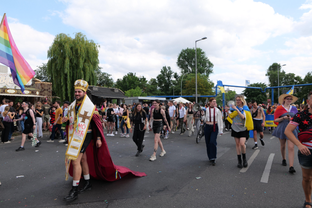 Eine Gruppe von Menschen marschiert bei der Pride Parade 2018 mit einer Regenbogenfahne und Musikinstrumenten, umgeben von Laternenmasten, Bäumen, Hütten und Wolken.