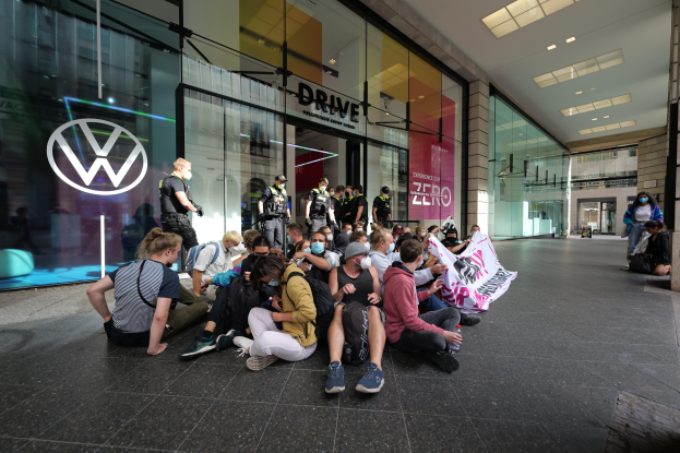 Gruppe von Menschen, die vor einem Volkswagen-Händler auf dem Boden sitzen und eine Protestfahne halten.
