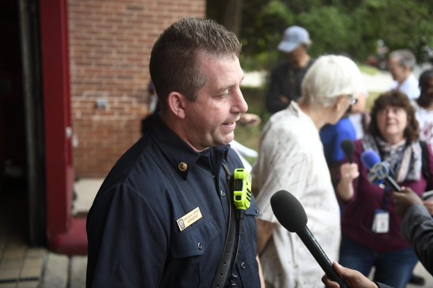 Polizeibeamter spricht zu einer Gruppe von Reportern mit Mikrofonen außerhalb bei Bäumen und einem Gebäude.