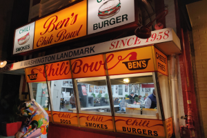 Ein Gebäude mit Glaswänden und beleuchteter Beschriftung, das ein paar Menschen im Inneren und eine Wand mit Rohren auf der rechten Seite zeigt.