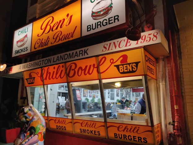 Ein Gebäude mit Glaswänden und beleuchteter Beschriftung, das ein paar Menschen im Inneren und eine Wand mit Rohren auf der rechten Seite zeigt.