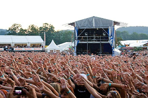 Eine große Menschenmenge auf einem Musikfestival mit erhobenen Händen, einige halten Mobiltelefone, mit einer Bühne, weißen Zelten und Bäumen im Hintergrund unter einem klaren blauen Himmel.