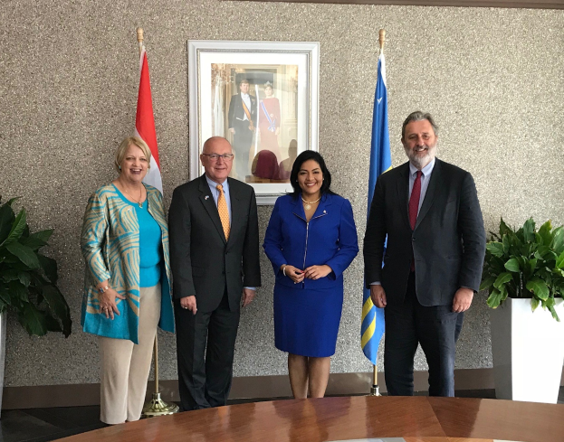 Eine Gruppe von Menschen, die um einen Tisch mit Papieren stehen und lächeln, umgeben von Fahnen, Topfpflanzen und einem Rahmenfoto an der Wand.