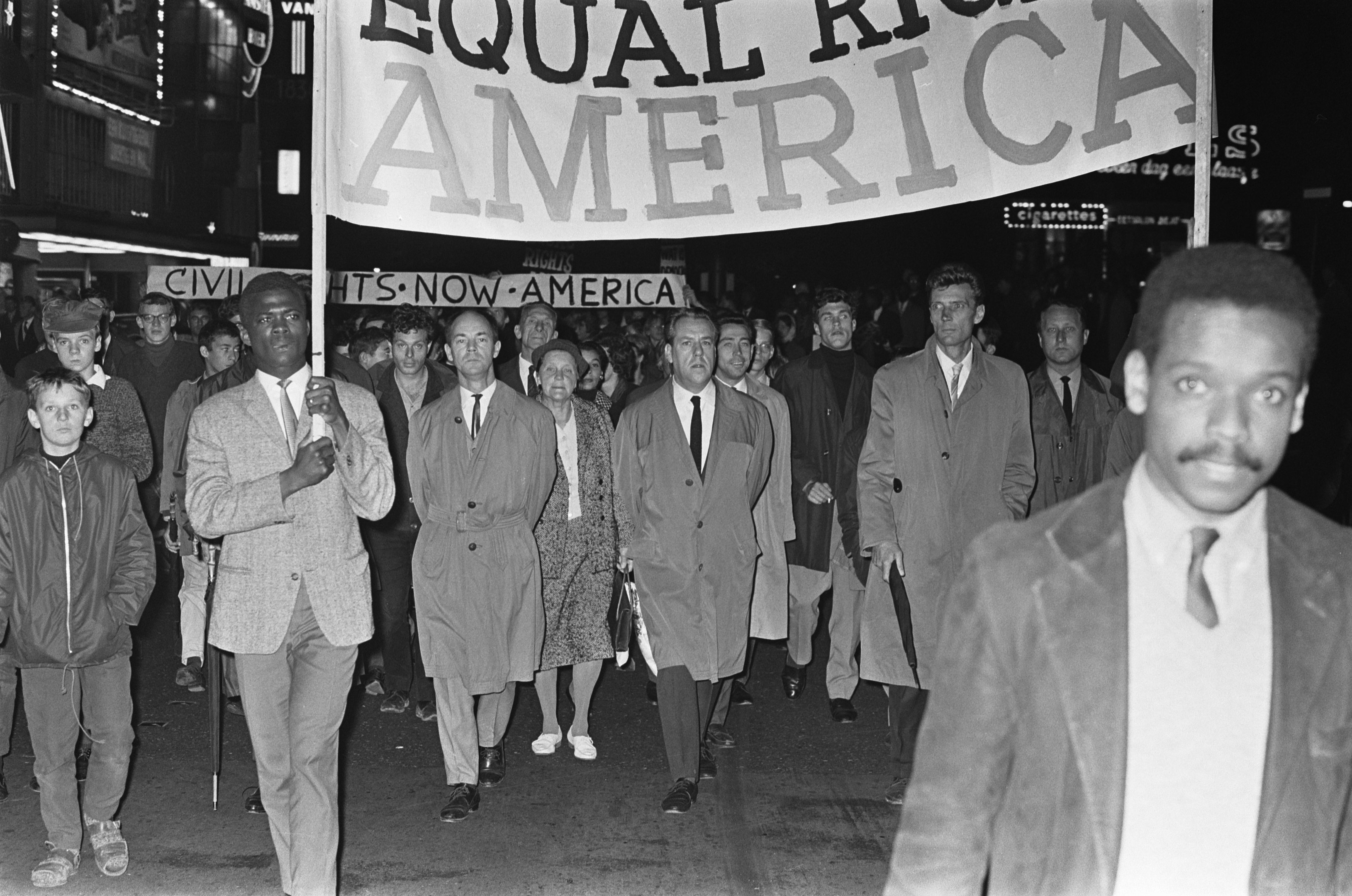 Schwarz-weiß-Foto einer Gruppe von Menschen, die mit einem Banner "Gleiche Rechte Amerika" die Straße entlang marschieren, mit Gebäuden und Straßenlaternen am Straßenrand.