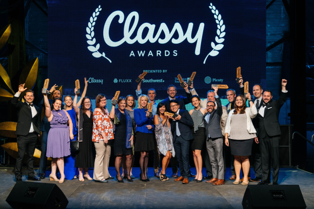 Eine Gruppe von Menschen auf einer Bühne, die strahlen und Auszeichnungen halten, mit Lautsprechern unten und einem Hintergrundbild, das "Classy Awards 2019" anzeigt.