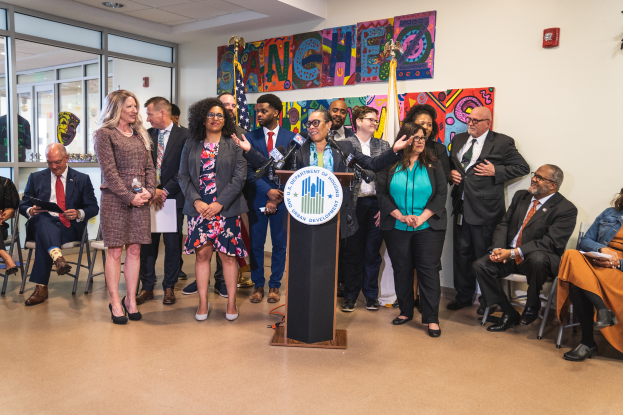 Eine Gruppe von Menschen, die vor einem Podium mit Mikrofon und einer Tafel mit der Aufschrift "Sancho City Councilman Joins City Council" steht und sitzt, mit Flaggen, Feuerlöscher und Deckenleuchten an der Rückwand.