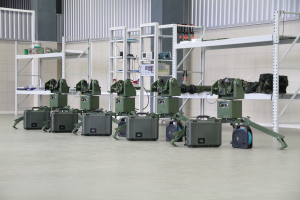 Gruppe von Milit├Ąrrobotern auf einem Boden mit Koffern und Objekten auf Gestellen, positioniert gegen eine Wand mit Gel├Ąnder im Hintergrund.