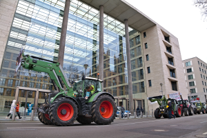 Eine Gruppe von Traktoren fährt eine Straße entlang vor einem Gebäude, mit Menschen auf dem Gehweg und einem Baum auf der rechten Seite, wahrscheinlich an einer Demonstration teilnehmend.