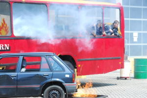 Roter Doppeldeckerbus mit Rauch und drei sichtbaren Passagieren, neben einem Auto geparkt, vor einem Glasfenster-Gebäude und einem Fass.