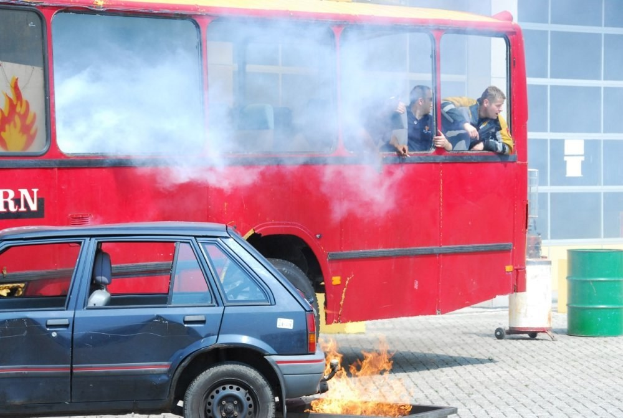 Roter Doppeldeckerbus mit Rauch und drei sichtbaren Passagieren, neben einem Auto geparkt, vor einem Glasfenster-Gebäude und einem Fass.