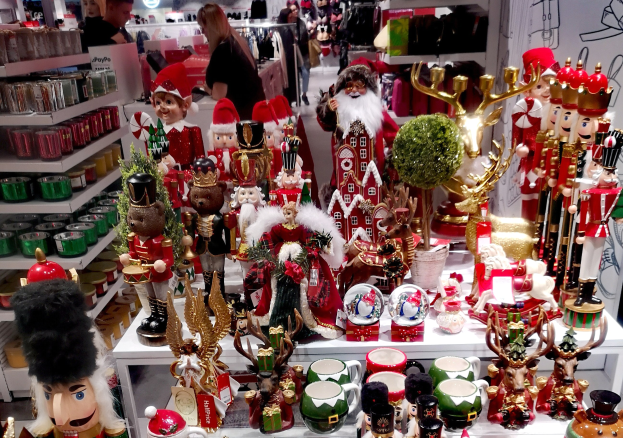 Ladeninneres mit einer Auswahl an Weihnachtsdekorationen, darunter Spielzeug, Tassen und andere Gegenstände auf Tischen und Regalen, mit Menschen, Lichtern und einer Wand im Hintergrund.