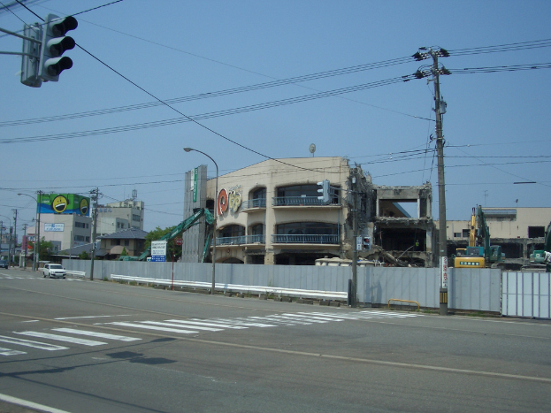 Eine Stadtstraße mit fahrenden Fahrzeugen, Strommasten, einer Ampel auf der linken Seite, Gebäuden, Bäumen und dem Himmel im Hintergrund sowie einem abbruchreifen Gebäude im Vordergrund.