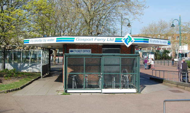 Haltestelle mit 'Gosport Ferry Ltd'-Schild, Metallzaun, Menschen, Fahrräder, Bäume, Straßenlaternen, Schilder, ein Gebäude mit Fenstern und eine bewölkte Himmel.