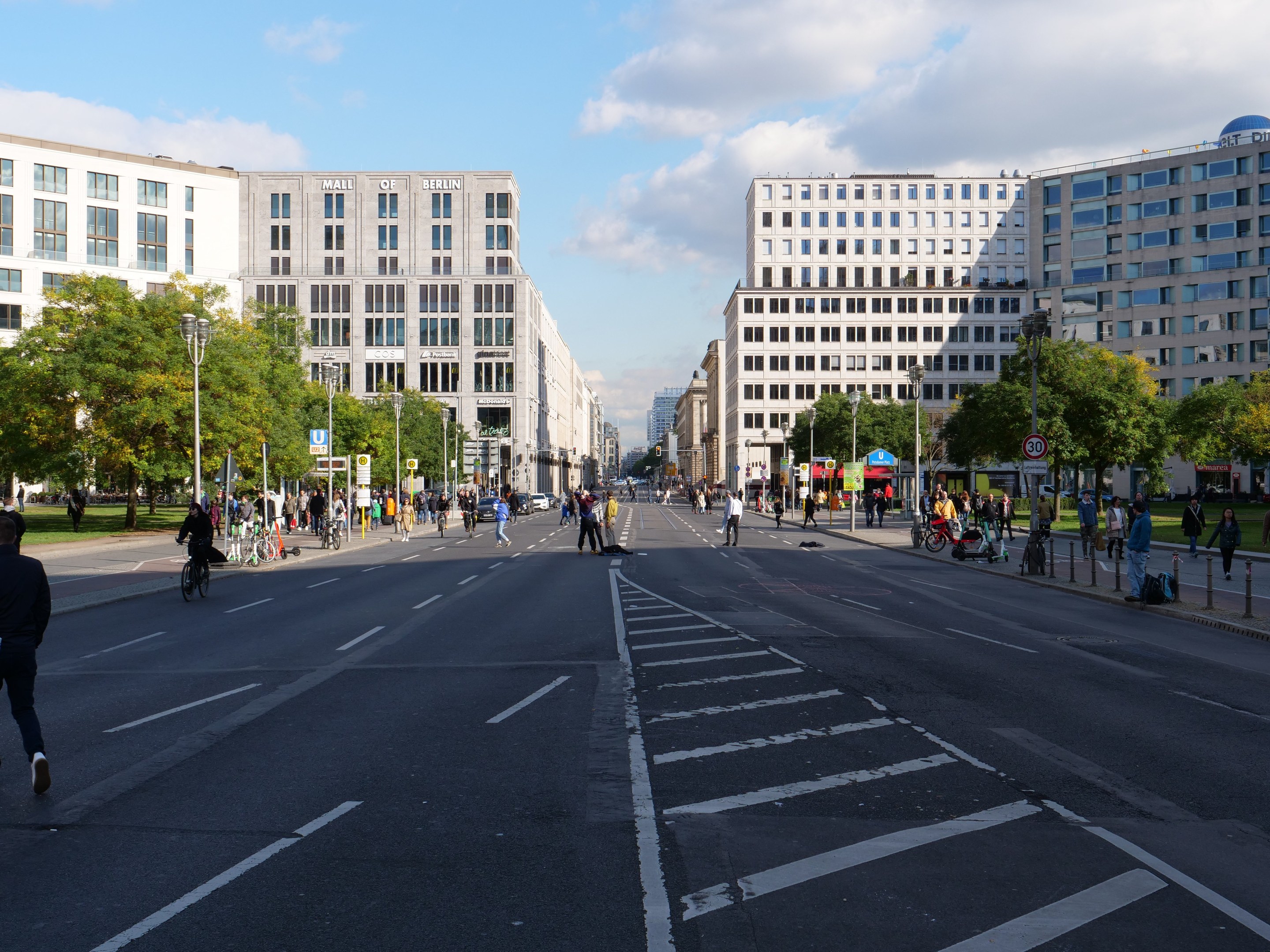 Eine belebte Stadtstraße in Berlin, Deutschland, mit Fußgängern und Radfahrern auf der Straße, hohen Gebäuden mit Fenstern, Bäumen, Laternen und Schildern unter einem bewölkten Himmel.