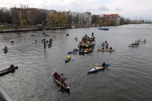 Eine Gruppe von Menschen in Kanus, die auf einem Fluss paddeln, mit einem Geländer auf der linken Seite, Bäumen, Gebäuden, Fahrzeugen und Pfählen im Hintergrund unter einem klaren blauen Himmel.