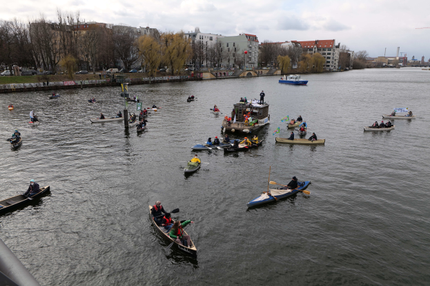 Eine Gruppe von Menschen in Kanus, die auf einem Fluss paddeln, mit einem Geländer auf der linken Seite, Bäumen, Gebäuden, Fahrzeugen und Pfählen im Hintergrund unter einem klaren blauen Himmel.