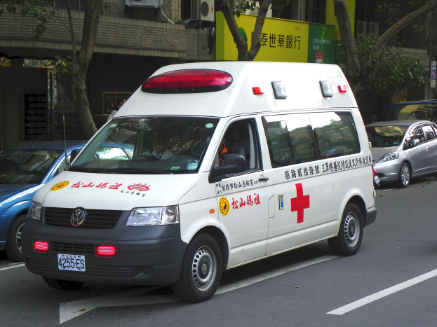 Ambulanz am Straßenrand mit anderen Fahrzeugen, Bäumen, Gebäuden, Texttafeln und Klimaanlagen im Hintergrund.