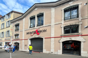 Eine Gruppe von Menschen mit Fahnen geht vor einem Gebäude mit einer "Montbron"-Tafel, einem Straßenschild und einer Banner mit Bildern und Text unter einem bewölkten Himmel die Straße entlang.