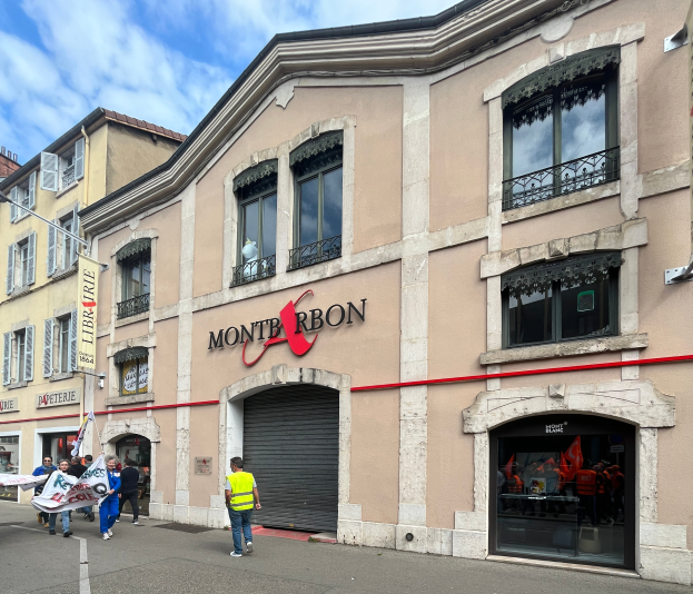 Eine Gruppe von Menschen mit Fahnen geht vor einem Gebäude mit einer "Montbron"-Tafel, einem Straßenschild und einer Banner mit Bildern und Text unter einem bewölkten Himmel die Straße entlang.