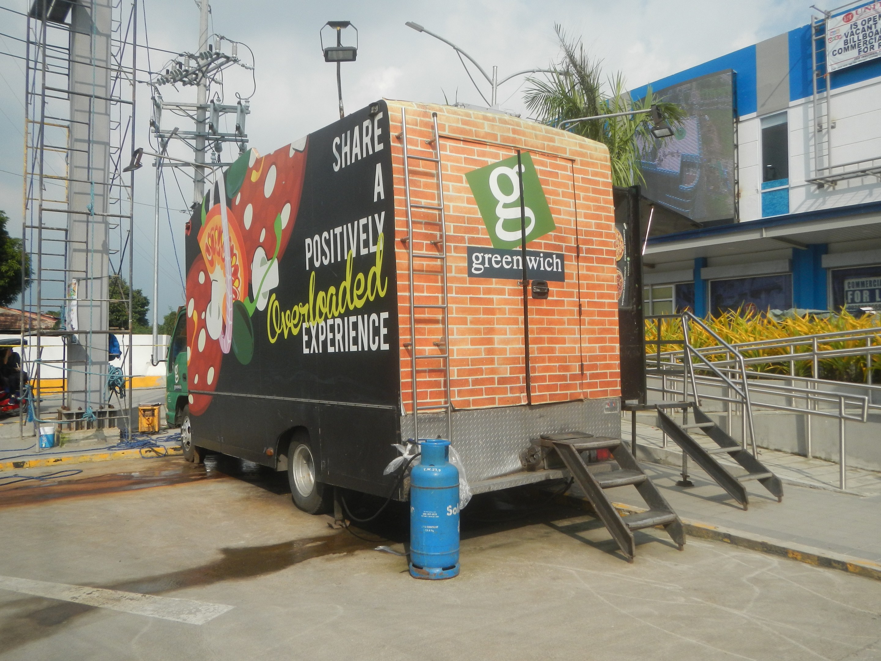 Ein Lkw parkt vor einem Gebäude mit einer Seite, auf der ein Schild mit der Aufschrift "Teilen Sie ein positiv überladenes Erlebnis" steht, mit einer Treppe, einem Zylinder auf dem Boden, Geländern, Strommasten mit Drähten, Laternen, Bäumen und einem bewölkten Himmel im Hintergrund.