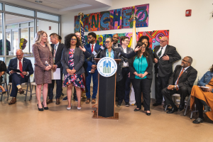 Gruppe von Menschen vor einem Rednerpult mit einem Mikrofon und einer Tafel mit der Aufschrift "Sancho-Stadtrat tritt dem Stadtrat bei", mit sitzenden und stehenden Teilnehmern, Flaggen, einem Feuerlöscher und Deckenleuchten.