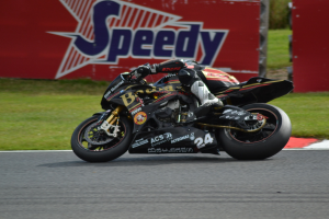 Eine Person in Helm und Schutzkleidung fährt ein Motorrad auf einer Rennstrecke, umgeben von Gras und einem Schild im Hintergrund.